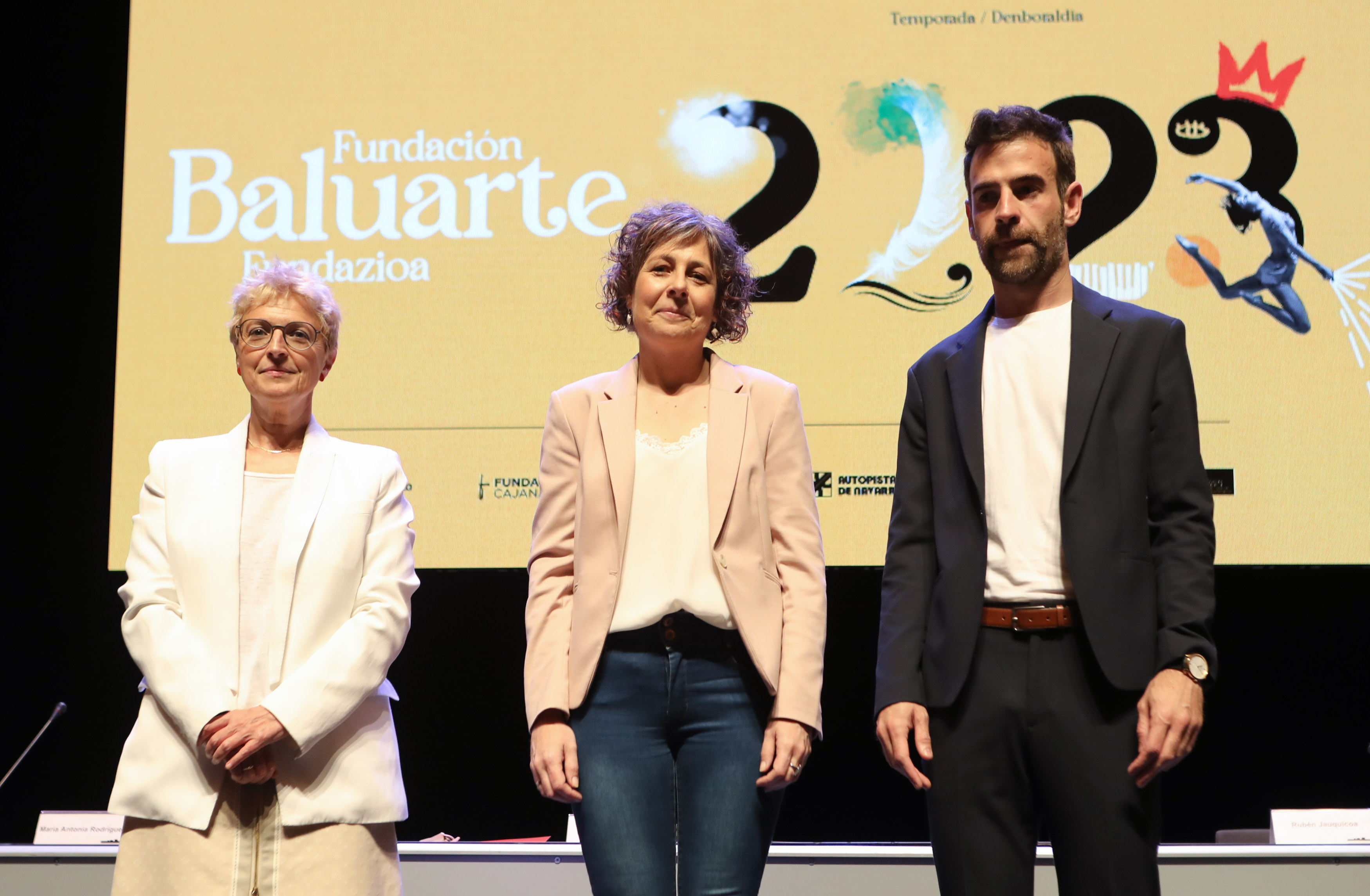 Fotografía de tres personas de pie y detrás el cartel promocional de la programación