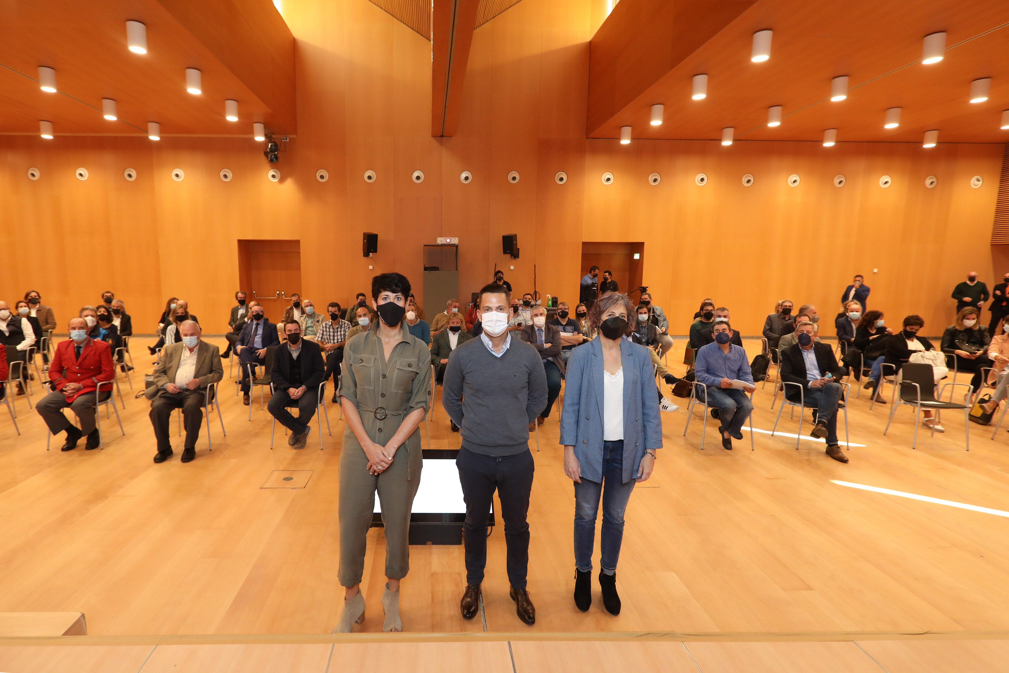 Fotografía de una sala con gente sentada y tres personas de pie.