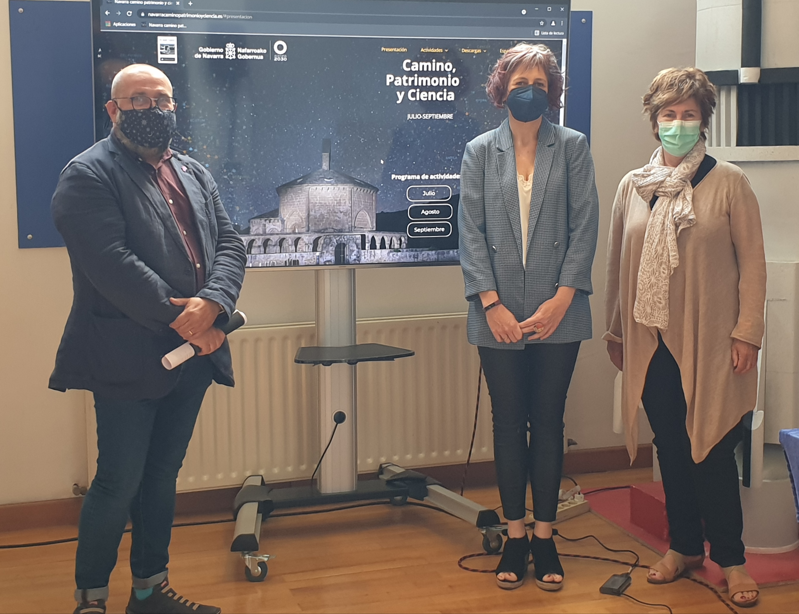 Fotografía de un hombre y dos mujeres de pie delante del cartel promocional