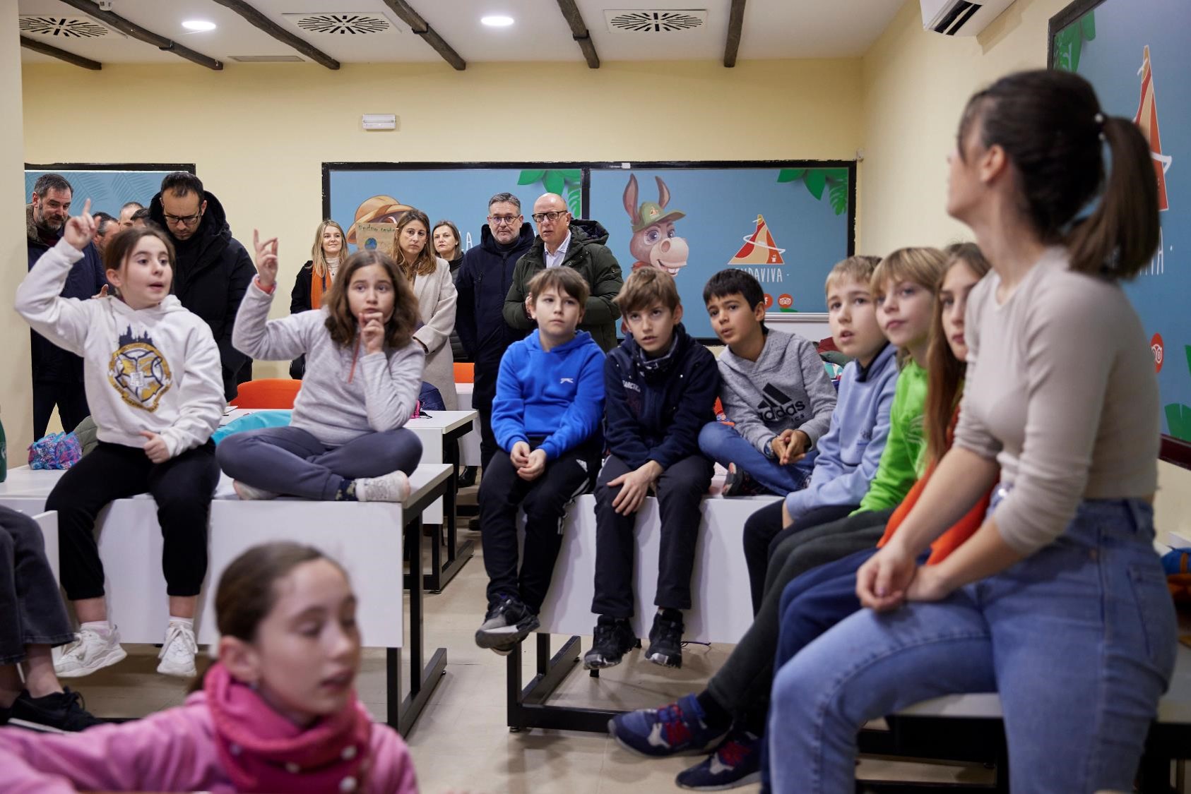 Imagen de un aula repleta de niños y niñas atendiendo a su profesora, algunos de ellos con la mano en alto. Al fondo, se ve al consejero de Educación, Carlos Gimeno, y al director gerente de CNAI, Juan Luis García.