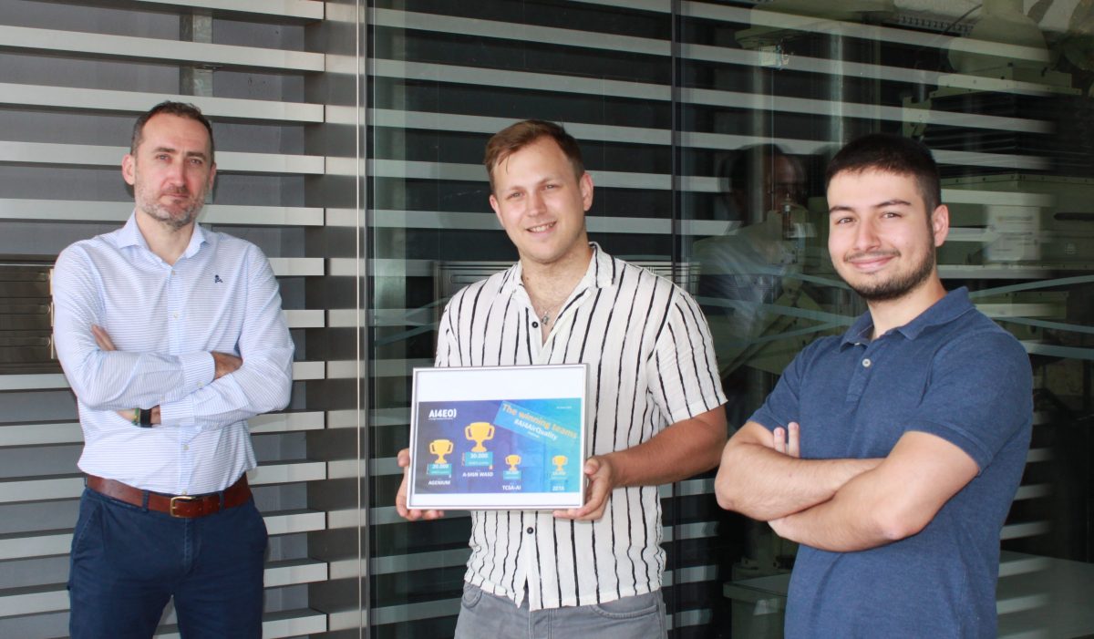 Fotografía de tres hombres de pie, uno de ellos con el premio en la mano