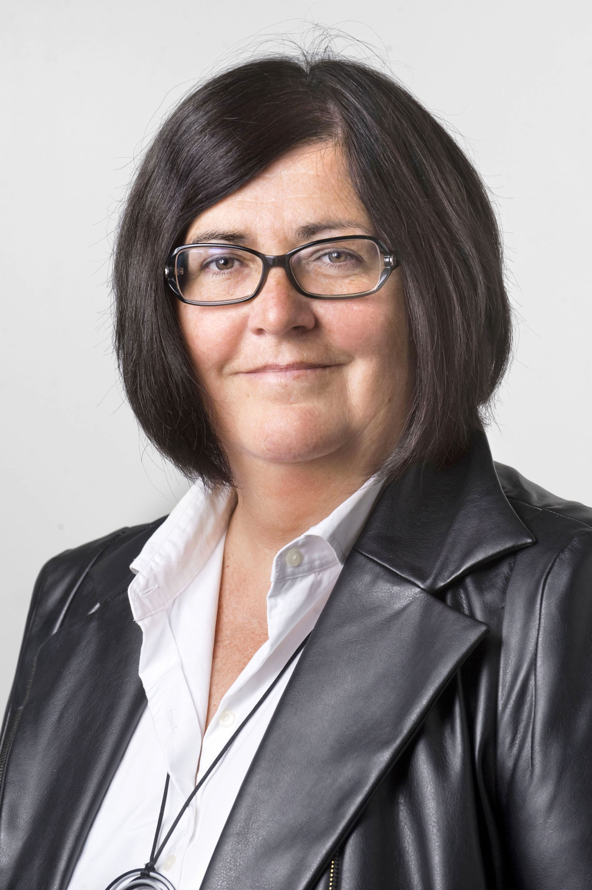 Fotografía de plano medio corto de Pilar Irogien, sonriente y ataviada con una cazadora de cuero negra con solapas, camisa blanca y gafas con montura negra. 