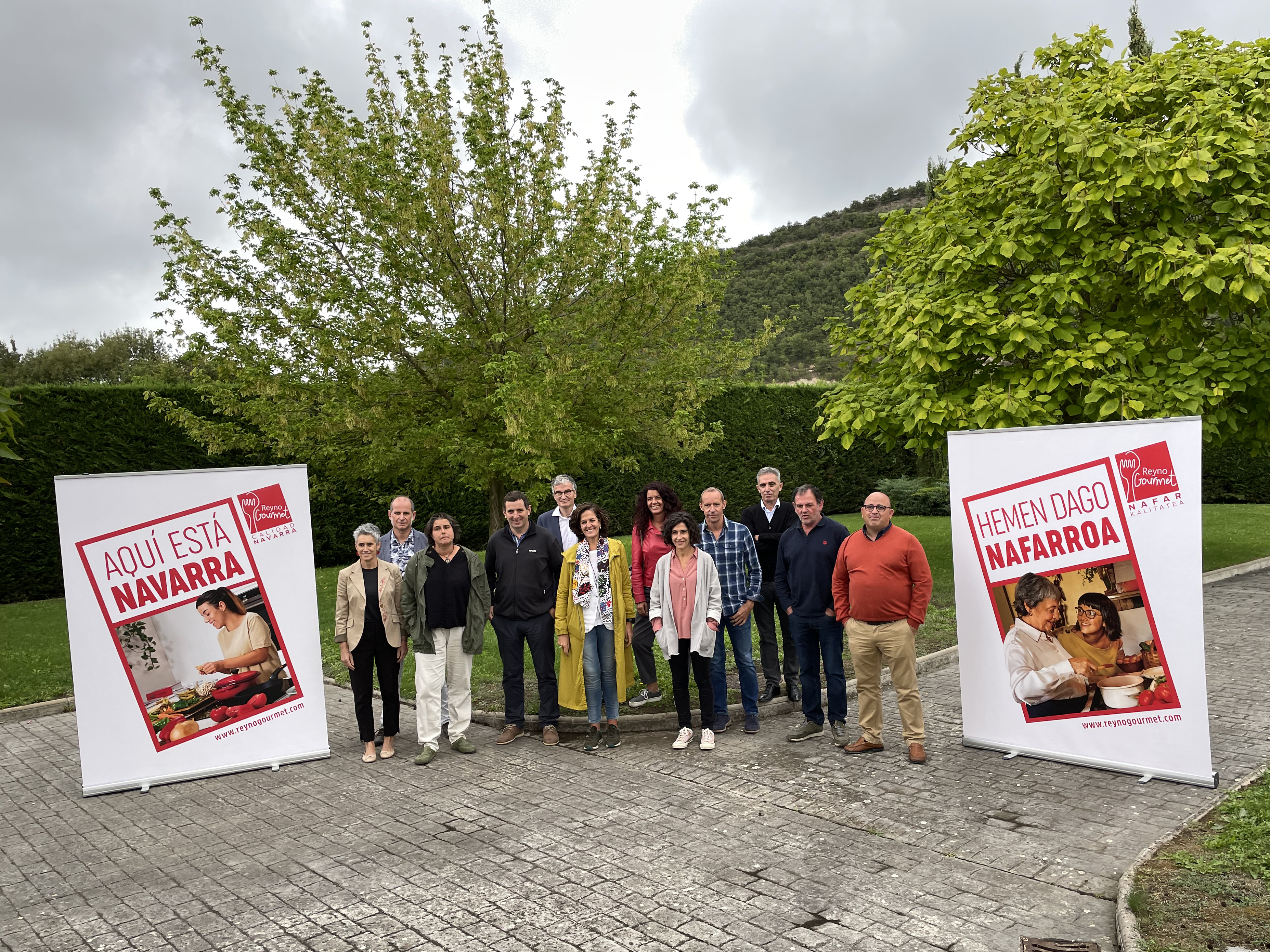 Fotografía de exterior de varias personas de pie y en cada lado el cartel promocional