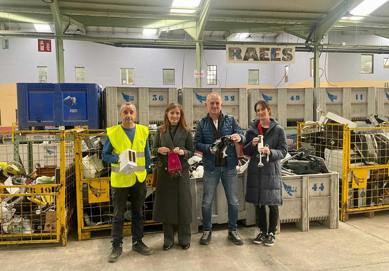 Fotografía de cuatro personas, dos hombres y dos mujeres, de cuerpo entero mostrando aparatos reciclados a cámara