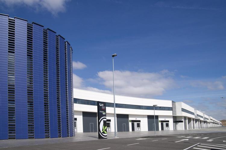 Panorámica de la fachada exterior del Vivero de Innovación Agroalimentario. A mano izquierda, se ve un edificio con forma cilíndrica de color azul. A continuación hacia la derecha, se extienden una larga fila de naves industriales bajas de color blanco.