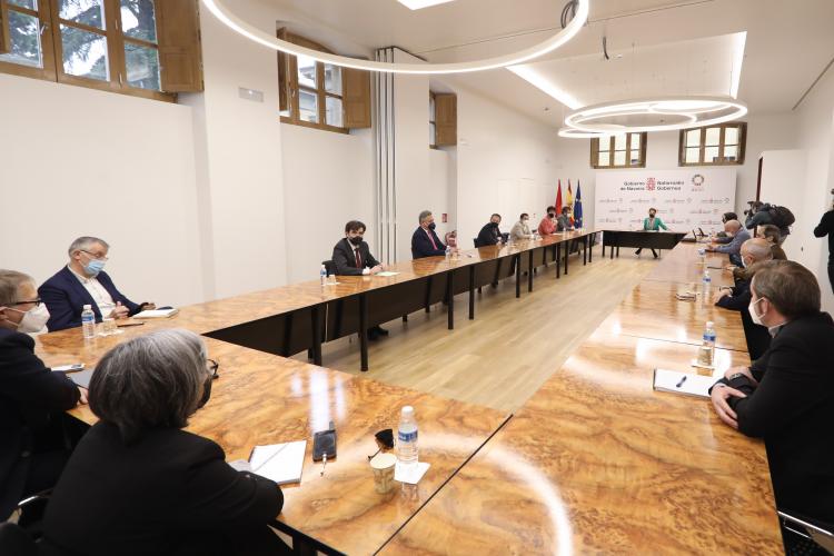 Fotografía de la conferencia en una sala, con una mesa grande y varias personas sentadas.