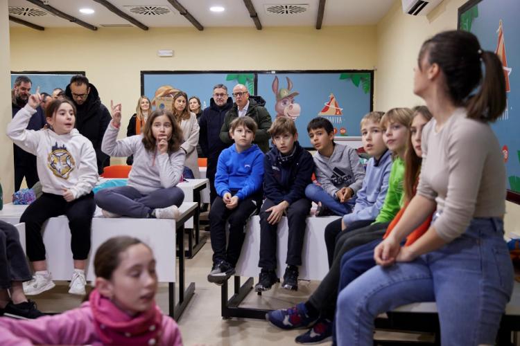 Imagen de un aula repleta de niños y niñas atendiendo a su profesora, algunos de ellos con la mano en alto. Al fondo, se ve al consejero de Educación, Carlos Gimeno, y al director gerente de CNAI, Juan Luis García.