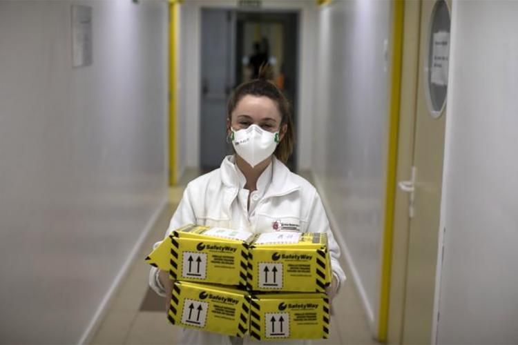 Fotografía interior de una enfermera con bata y mascarilla blancas llevando cuatro cajas amarillas