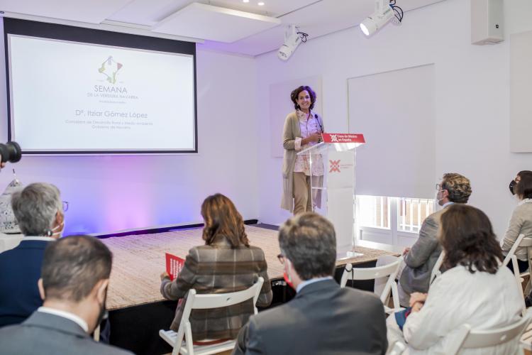 Fotografía de la consejera de pie en un atril durante la presentación y varias personas sentadas.