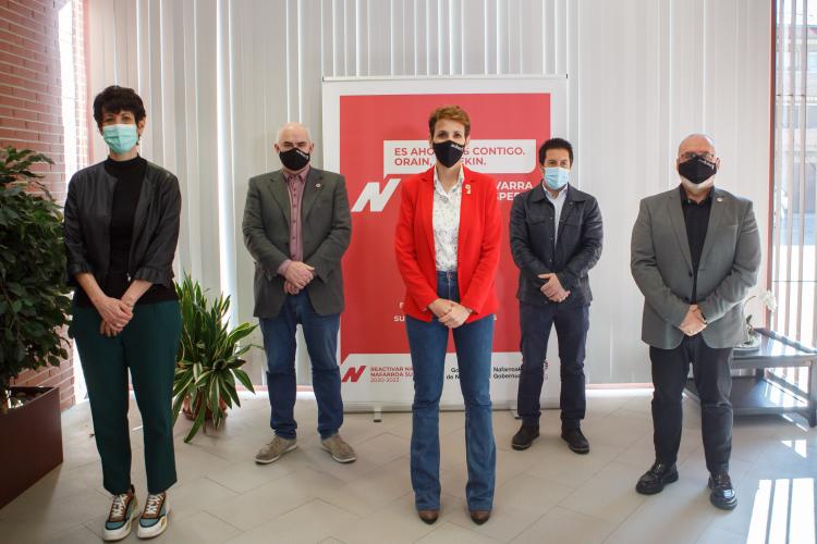 Fotografía interior donde aparecen dos mujeres y tres hombres de pie con un cartel promocional de color rojo detrás de ellos.