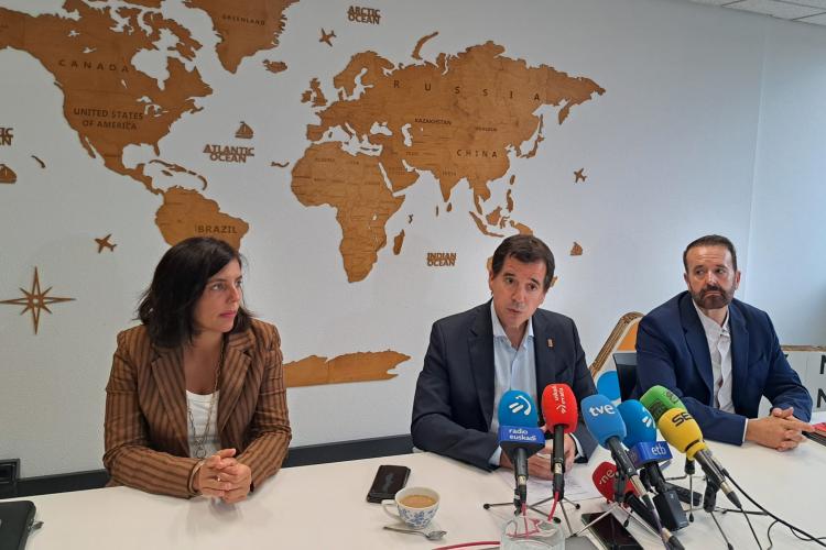 3 personas sentados en una mesa delante de micrófonos de prensa
