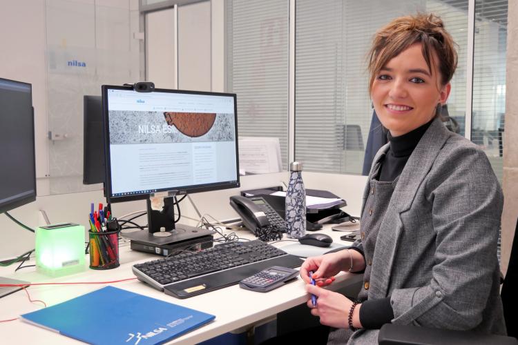 Fotografía de un puesto de trabajo. Amaia, sonriente, posa sentada en su mesa delante del ordenador. 
